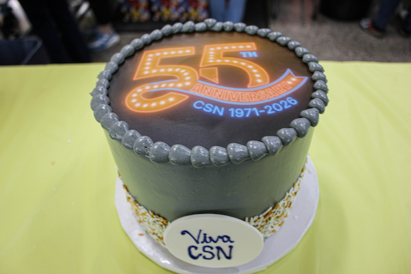 A gray cake with a "55th Anniversary CSN 1971-2026" design and "Viva CSN" plaque, displayed on a yellow table.