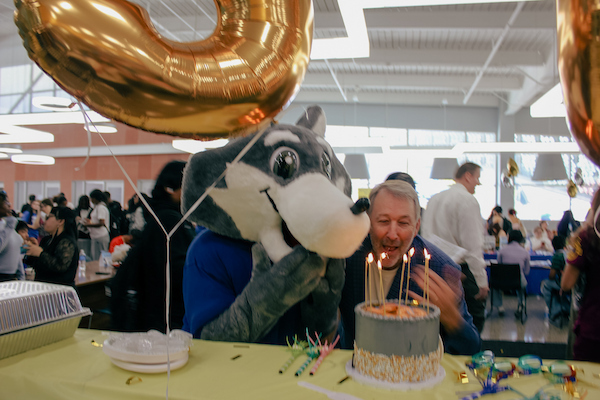 A person in a raccoon mascot costume blows out candles on a birthday cake while another person watches, with a large gold balloon nearby.