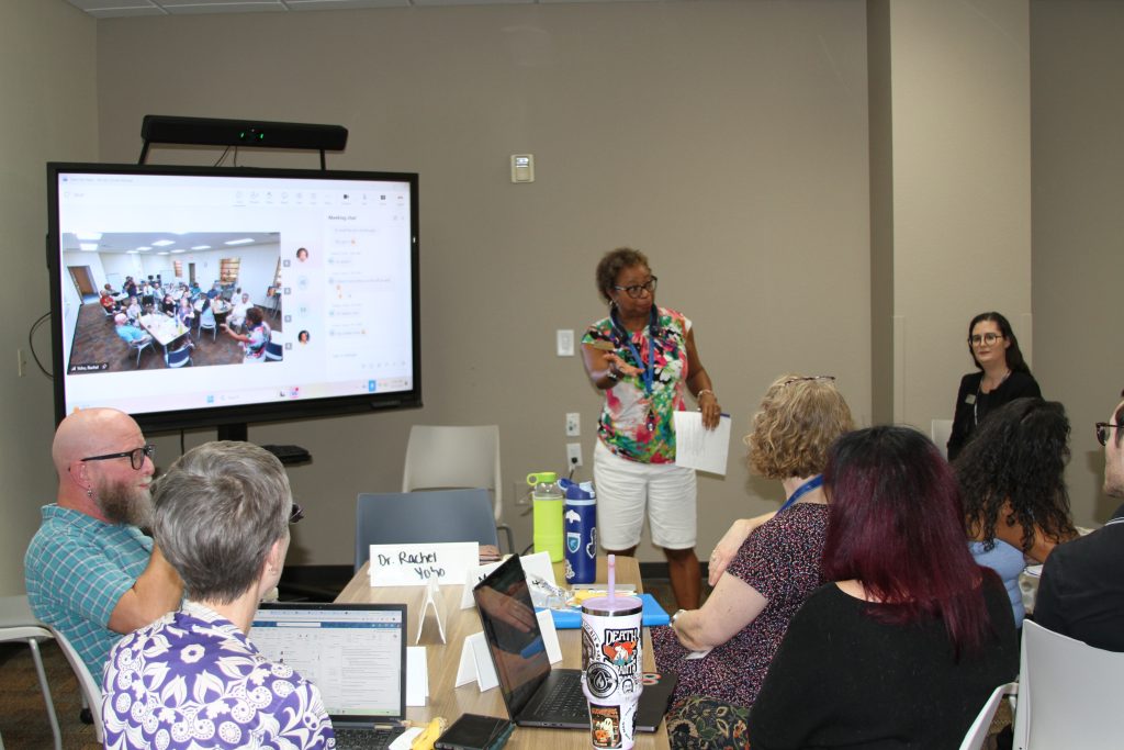 A presenter stands before an audience in a conference room, addressing attendees while a large screen displays a live video feed of the same meeting.