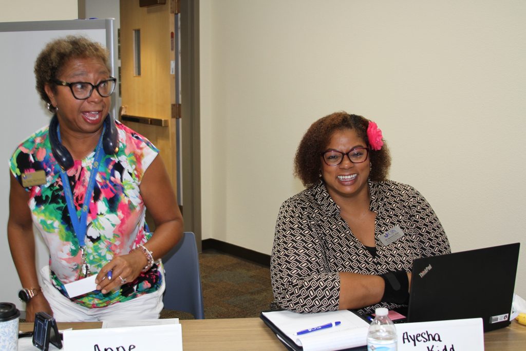 Two individuals are seated at a table in a meeting setting, one smiling while using a laptop and the other standing with headphones around their neck.