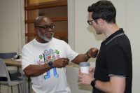 Two individuals engage in conversation indoors, one holding a coffee cup and the other gesturing while wearing a colorful graphic tee.