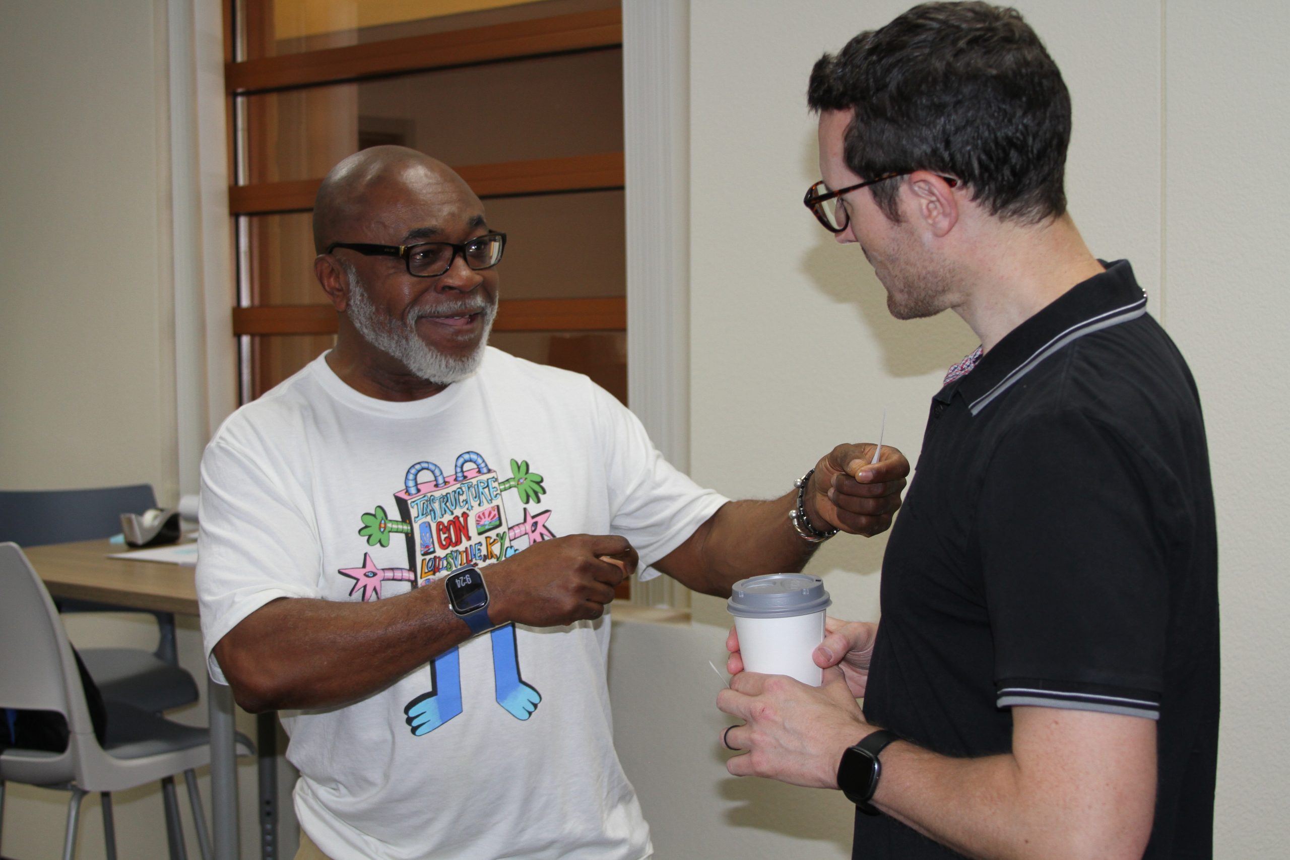Two individuals engage in conversation indoors, one holding a coffee cup and the other gesturing while wearing a colorful graphic tee.