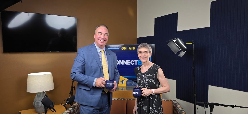 Two individuals stand in a studio setting, holding mugs with “CSN” branding, in front of a screen displaying “ON AIR” and “CONNECTED.”