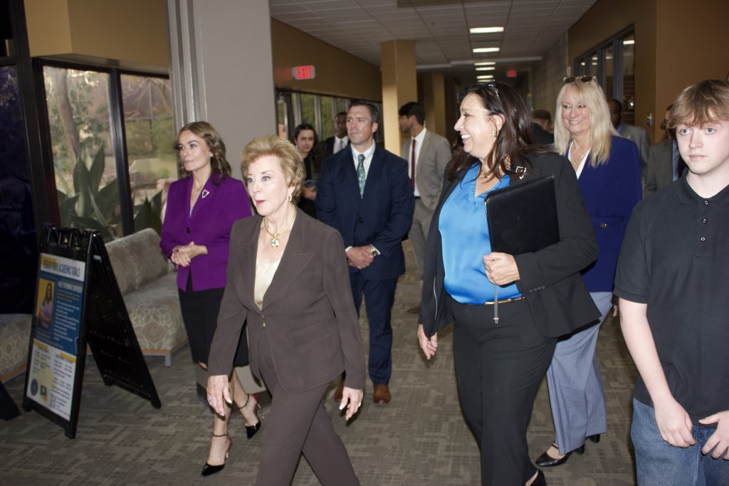 A group of people, including a woman in a brown suit and another in a blue blouse holding a folder, walk through a hallway in an office building.