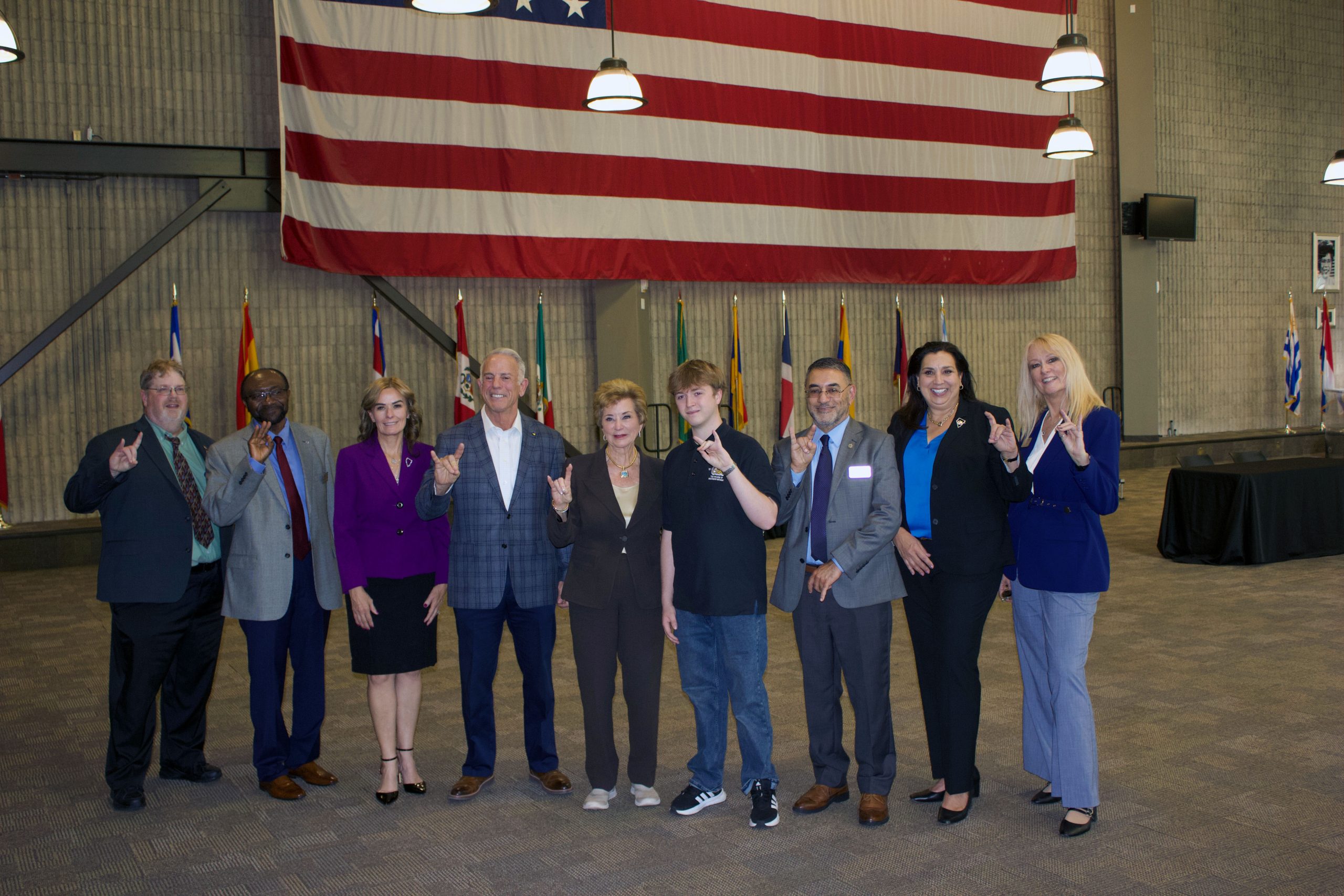 A group of nine individuals pose together indoors in front of a large American flag, making hand gestures while dressed in formal or business attire.