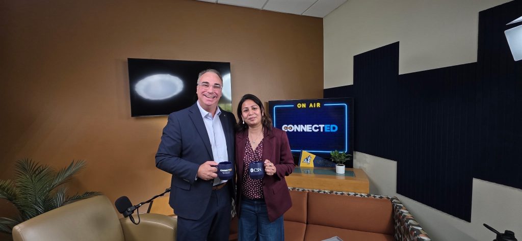 Two individuals stand in a studio setting, smiling for the camera, with one holding a branded mug and a screen displaying "ON AIR" and "CONNECTED" behind them.