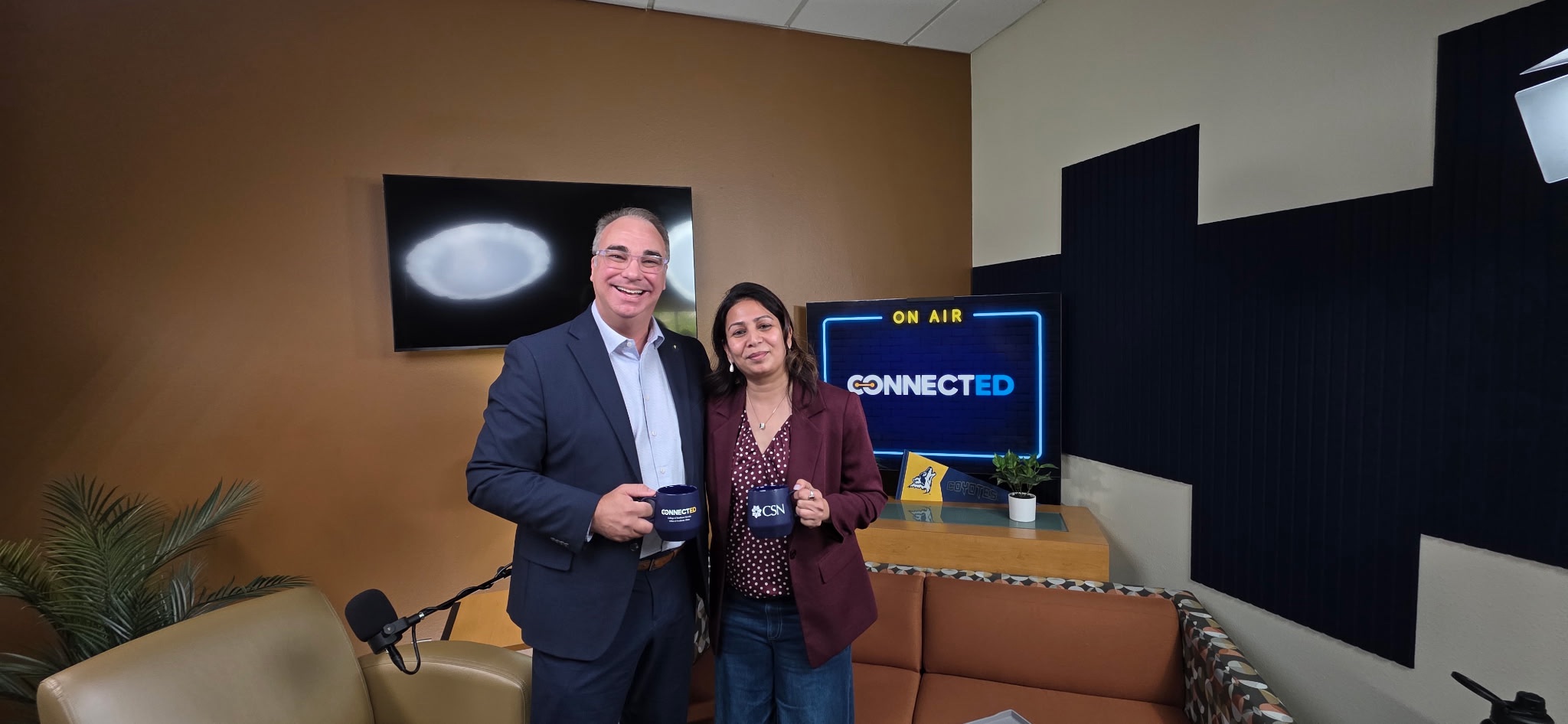 Two individuals stand in a studio setting, smiling for the camera, with one holding a branded mug and a screen displaying "ON AIR" and "CONNECTED" behind them.