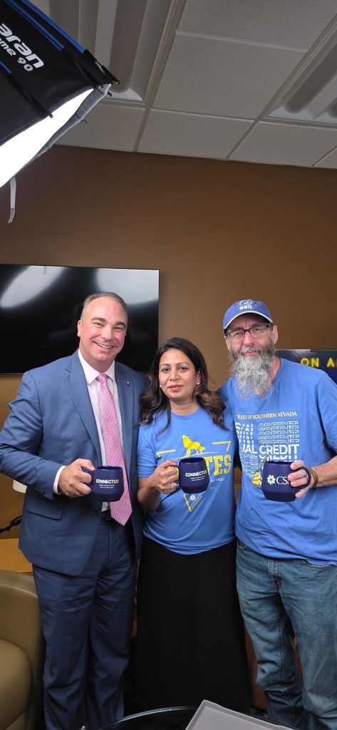 Three individuals pose together indoors, each holding a branded mug, with one person wearing a suit and tie, another in a blue t-shirt with a logo, and the third in a blue t-shirt and cap.